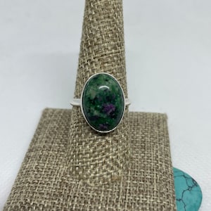 May include: A silver ring with a green and purple oval gemstone. The ring is on a brown fabric ring holder.
