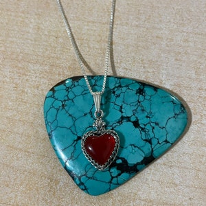 May include: A silver chain necklace with a red heart pendant. The heart is set in a silver frame with a detailed design. The necklace is displayed on a turquoise stone.