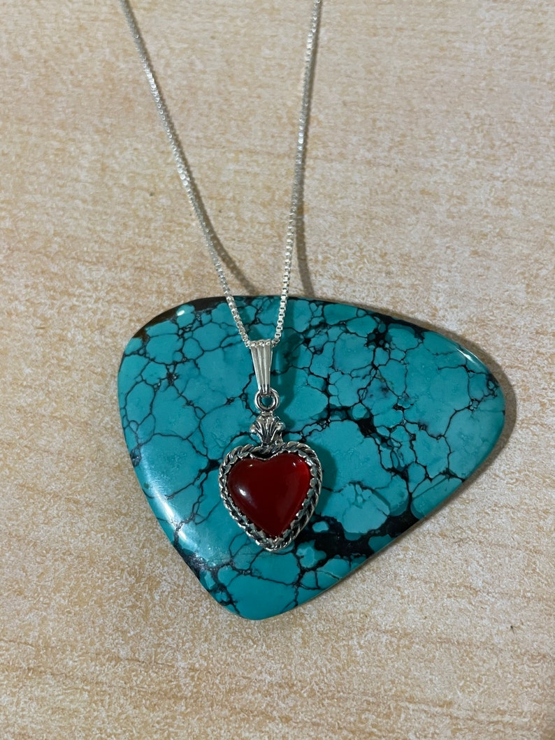 May include: A silver chain necklace with a red heart pendant. The heart is set in a silver frame with a detailed design. The necklace is displayed on a turquoise stone.