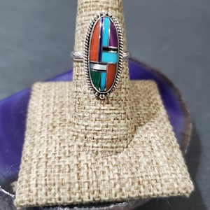 Op de afbeelding: Een zilveren ring met een ovale steen die in het midden is gezet. De steen is gemaakt van verschillende kleuren turquoise, koraal en andere stenen. De ring staat op een displaystandaard.