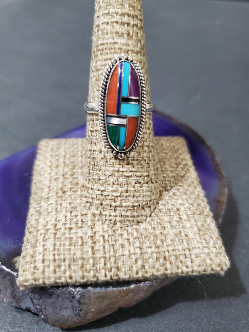 Op de afbeelding: Een zilveren ring met een ovale steen die in het midden is gezet. De steen is gemaakt van verschillende kleuren turquoise, koraal en andere stenen. De ring staat op een displaystandaard.