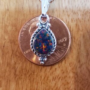 May include: A silver necklace with a teardrop-shaped opal pendant set in a silver bezel. The pendant is surrounded by a silver rope design. The necklace is shown next to a US penny for size comparison.