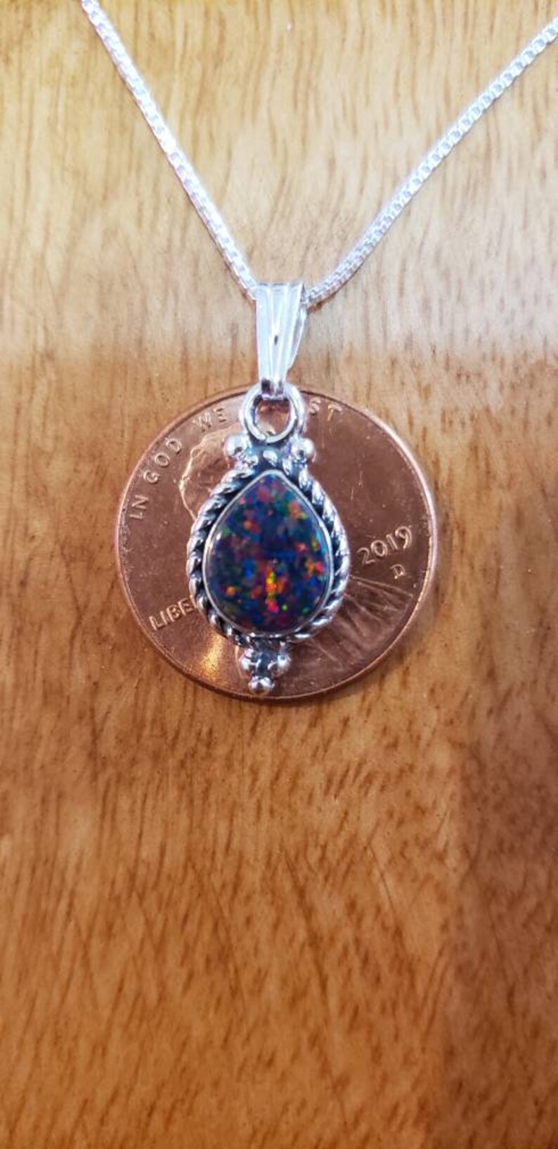 May include: A silver necklace with a teardrop-shaped opal pendant set in a silver bezel. The pendant is surrounded by a silver rope design. The necklace is shown next to a US penny for size comparison.