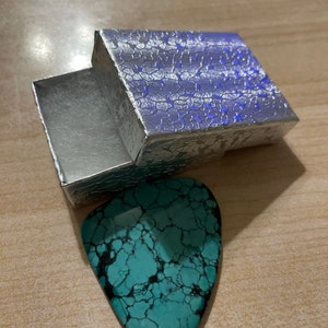 Puede incluir: Una p&uacute;a de guitarra azul turquesa con un patr&oacute;n de m&aacute;rmol en una caja plateada con superficie texturizada.