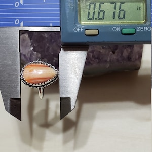 May include: A close-up of a ring with a teardrop-shaped, orange and white stone. The stone is 0.676 inches wide. The ring is set in silver.