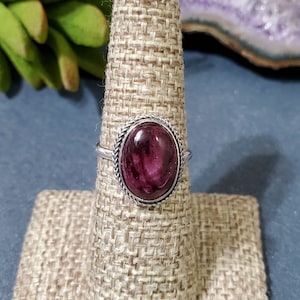 May include: A silver ring with a purple and pink oval gemstone set in a silver bezel. The ring is on a brown fabric ring display stand.