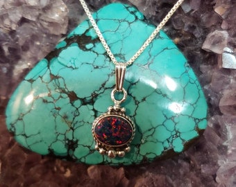 May include: A silver chain necklace with a dark red opal pendant set in a silver bezel. The pendant is surrounded by small silver beads. The necklace is displayed on a turquoise stone.