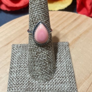 May include: A silver ring with a teardrop-shaped pink stone set in a silver bezel. The ring is on a ring display stand.