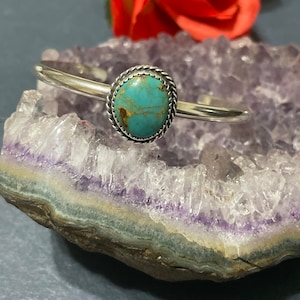 Puede incluir: Un brazalete de plata con una piedra turquesa engastada en un bisel de plata. La piedra tiene un patrón moteado de azul, verde y marrón.