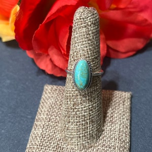 May include: A silver ring featuring an oval turquoise stone. The stone is set in a silver band with a twisted rope design. The ring is displayed on a burlap ring holder.
