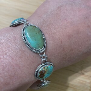 May include: Silver bracelet featuring oval turquoise stones. The stones are a light green-blue color, set in a silver frame with a twisted rope design around the edges. The bracelet is connected by silver links.