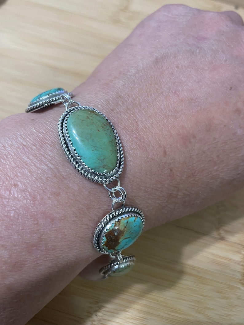 May include: Silver bracelet featuring oval turquoise stones. The stones are a light green-blue color, set in a silver frame with a twisted rope design around the edges. The bracelet is connected by silver links.