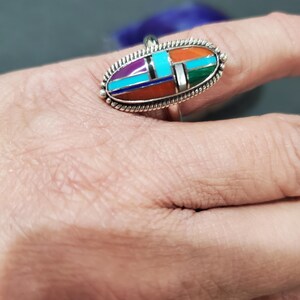 Op de afbeelding: Een zilveren ring met een ovale steen die in het midden is gezet. De steen is gemaakt van meerdere kleuren turquoise, koraal en andere stenen. De ring is gezet met een zilveren band.