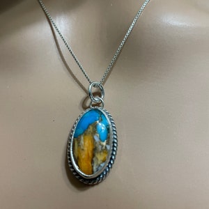 May include: A silver chain necklace with a pendant featuring a large oval-shaped turquoise stone with brown and white markings. The stone is set in a silver bezel with a rope-like border.