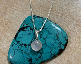 May include: A silver chain necklace with a round, white moonstone pendant. The necklace is displayed on a turquoise stone.