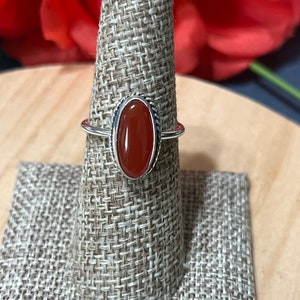 May include: A silver ring with an oval-shaped carnelian gemstone. The ring is set on a brown fabric ring holder.