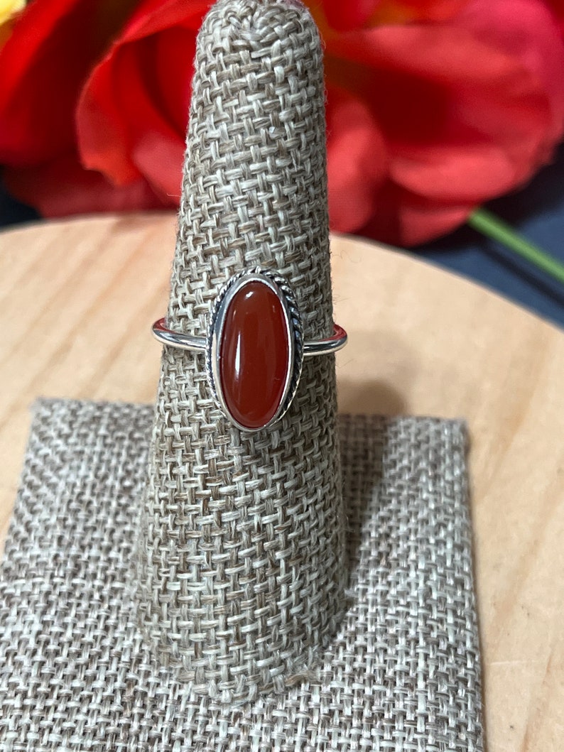 May include: A silver ring with an oval-shaped carnelian gemstone. The ring is set on a brown fabric ring holder.