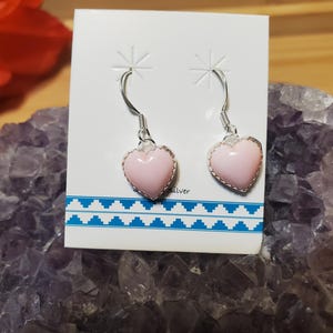 May include: A pair of silver earrings with pink heart-shaped pendants. The earrings are displayed on a white card with a blue geometric pattern. The word "silver" is printed on the card. The earrings are set against a purple crystal background.