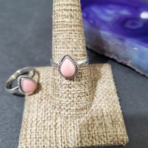 May include: Two silver rings with pink gemstone accents. The ring on the left is a simple band with a teardrop-shaped stone. The ring on the right has a more ornate setting with a teardrop-shaped stone.