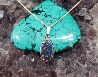 May include: A silver chain necklace with an oval-shaped opal pendant. The opal is a dark iridescent blue, green, and red. The necklace is displayed on a bed of purple amethyst crystals.