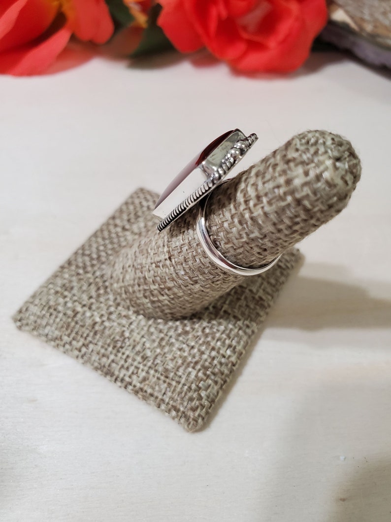 May include: A silver ring with a large, dark red gemstone set in a bezel setting. The ring is on a brown fabric ring display stand.