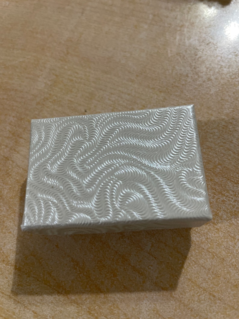 May include: A small, white, rectangular box with a swirling pattern on the surface. The box is made of cardboard and has a smooth finish.