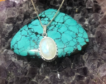 May include: A silver chain necklace with a white oval moonstone pendant. The pendant is set in a silver bezel and has a delicate, intricate design. The necklace is displayed on a bed of purple amethyst crystals.