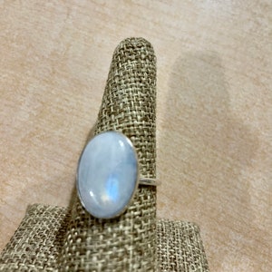 Puede incluir: Un anillo de plata con una gran piedra de luna ovalada. El anillo est&aacute; en un soporte de anillo de tela.
