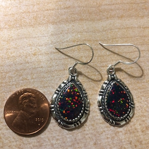 May include: A pair of silver dangle earrings with teardrop-shaped black opal stones. The stones have a red, orange, and green iridescent sheen. The earrings are about the size of a US penny.