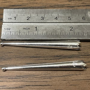 May include: Two silver metal knitting needles with pointed tips and rounded ends. The needles are approximately 2 inches long and are lying on a wooden surface.