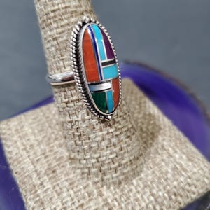 Op de afbeelding: Een zilveren ring met een ovale steen in het midden. De steen is gemaakt van turquoise, koraal en andere gekleurde stenen. De ring staat op een displaystandaard.