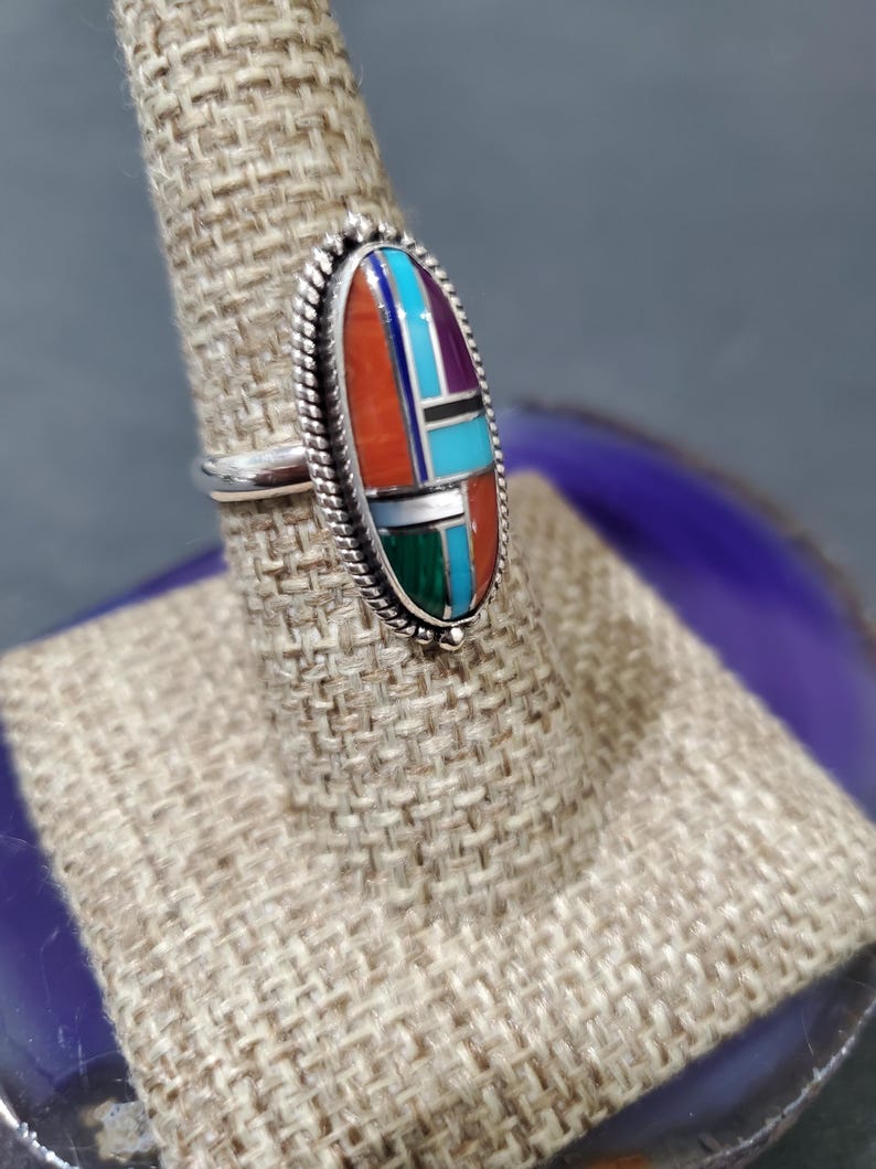 Op de afbeelding: Een zilveren ring met een ovale steen in het midden. De steen is gemaakt van turquoise, koraal en andere gekleurde stenen. De ring staat op een displaystandaard.
