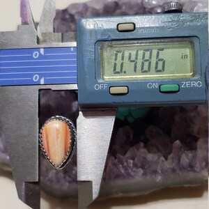 May include: A close-up of a teardrop-shaped, orange and white gemstone ring with a silver setting. The ring is being measured with a digital caliper, which reads 0.486 inches.