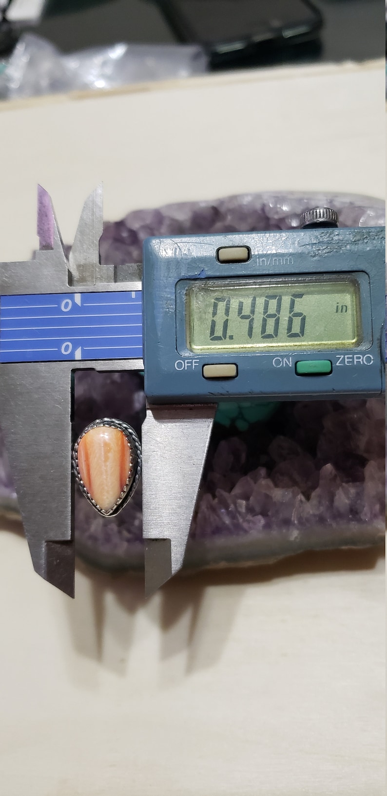 May include: A close-up of a teardrop-shaped, orange and white gemstone ring with a silver setting. The ring is being measured with a digital caliper, which reads 0.486 inches.