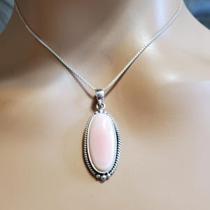 May include: A silver necklace with a pendant featuring a large, oval, light pink stone. The stone is set in a silver frame with a rope-like border and small silver beads. The chain is a delicate box chain.