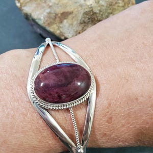 May include: A silver bangle bracelet with a large oval-shaped purple gemstone set in the center. The bracelet has a delicate, twisted wire design.