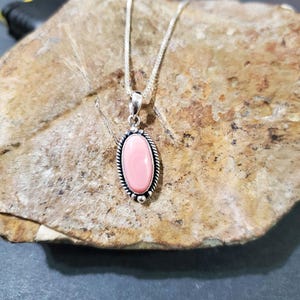 May include: A silver chain necklace with a pink oval pendant set in a silver frame. The pendant is on a rock background.