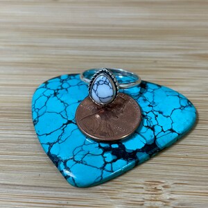 May include: A silver ring with a white turquoise stone set in a teardrop shape. The ring is on a turquoise background with a penny for scale.