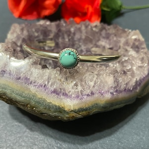 May include: A silver bangle bracelet with a turquoise stone set in a silver bezel. The bracelet is resting on a purple amethyst geode.