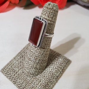 May include: A silver ring with a rectangular carnelian stone set in a bezel setting. The ring is on a fabric ring display stand.