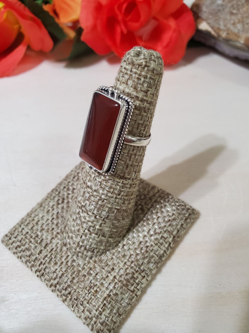 May include: A silver ring with a rectangular carnelian stone set in a bezel setting. The ring is on a fabric ring display stand.