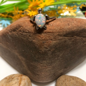 May include: A silver ring with a white opal turtle charm. The ring is on a brown rock.