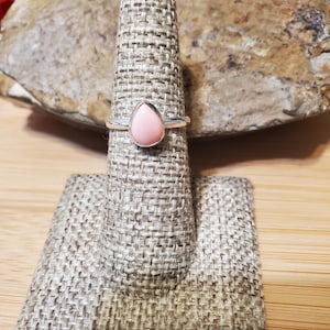 Puede incluir: Un anillo de plata con una piedra preciosa rosa pálido en forma de lágrima. El anillo se exhibe en un soporte para anillos beige texturizado. El fondo incluye una roca texturizada y una superficie de madera.