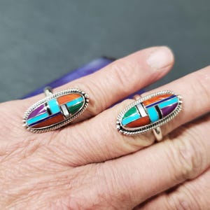 Op de afbeelding: Twee zilveren ringen met ovale inlegsels van turquoise, koraal en paarse steen. De ringen zitten aan een vinger.