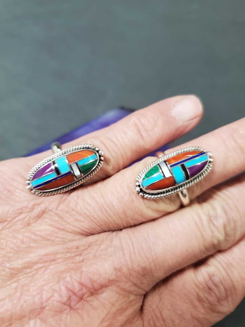 Op de afbeelding: Twee zilveren ringen met ovale inlegsels van turquoise, koraal en paarse steen. De ringen zitten aan een vinger.
