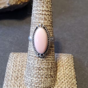 May include: A silver ring featuring a smooth, oval-shaped light pink stone set in a decorative twisted metal border. The band is polished silver, and the setting is framed by small silver beads, creating a detailed and classic jewelry piece.