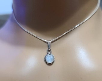 May include: A silver necklace with a small, round pendant. The pendant features a light-colored, possibly moonstone, center stone surrounded by a twisted metal border. The chain is a delicate, silver box chain.