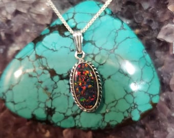 May include: A silver pendant necklace featuring an oval-shaped, black opal gemstone with vibrant red and orange flashes. The opal is set in a silver frame with a rope-like border, suspended from a delicate silver chain. The necklace rests on a turquoise stone.
