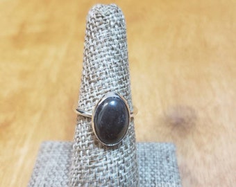 Puede incluir: Un anillo de plata con una piedra grande, ovalada, de color gris oscuro. El anillo se exhibe en un soporte de tela gris sobre un fondo de madera marrón claro. La piedra está engastada en una banda de plata pulida y sencilla.