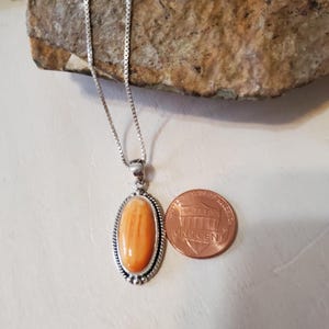 May include: Silver necklace with an oval, orange pendant. The pendant is set in a silver frame with a beaded edge. A U.S. one-cent coin is next to the pendant for size comparison. The necklace is on a light-colored surface.